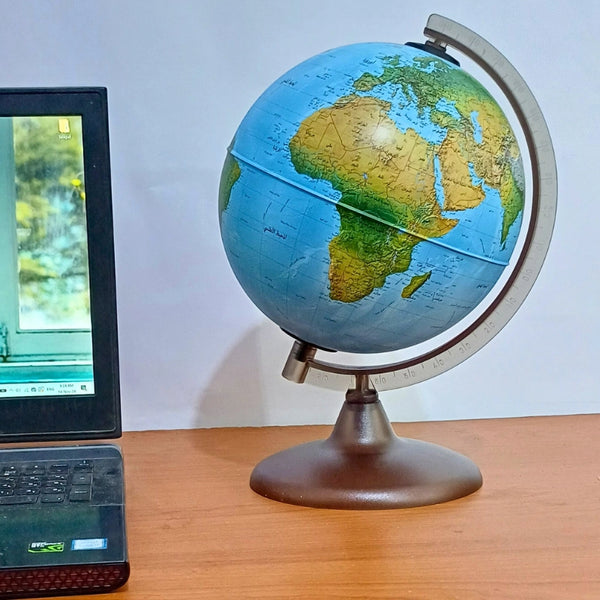 A 20 cm Arabic globe displayed on a wooden desk beside a laptop, with a focus on the African and European continents. The globe sits on a metallic base and features Arabic script for the country names